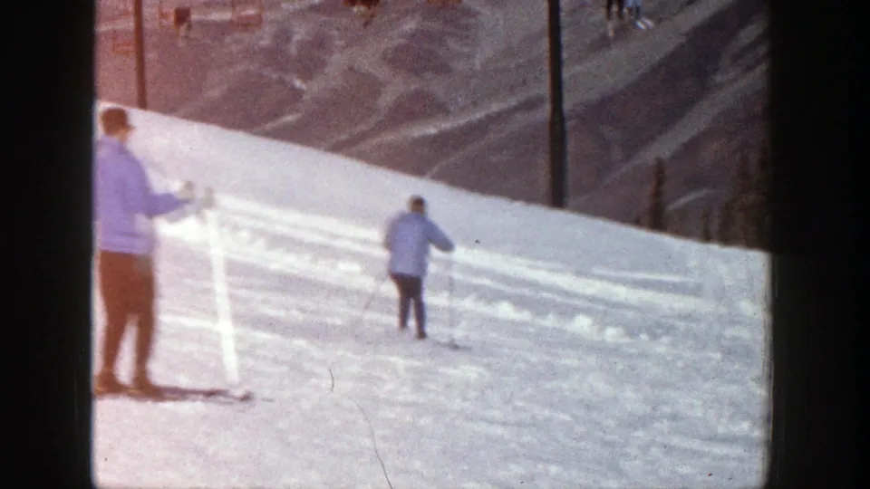 1961 Aspen home-movie frame showing two skiers descending a bright snowy slope with distant mountain terrain behind them.