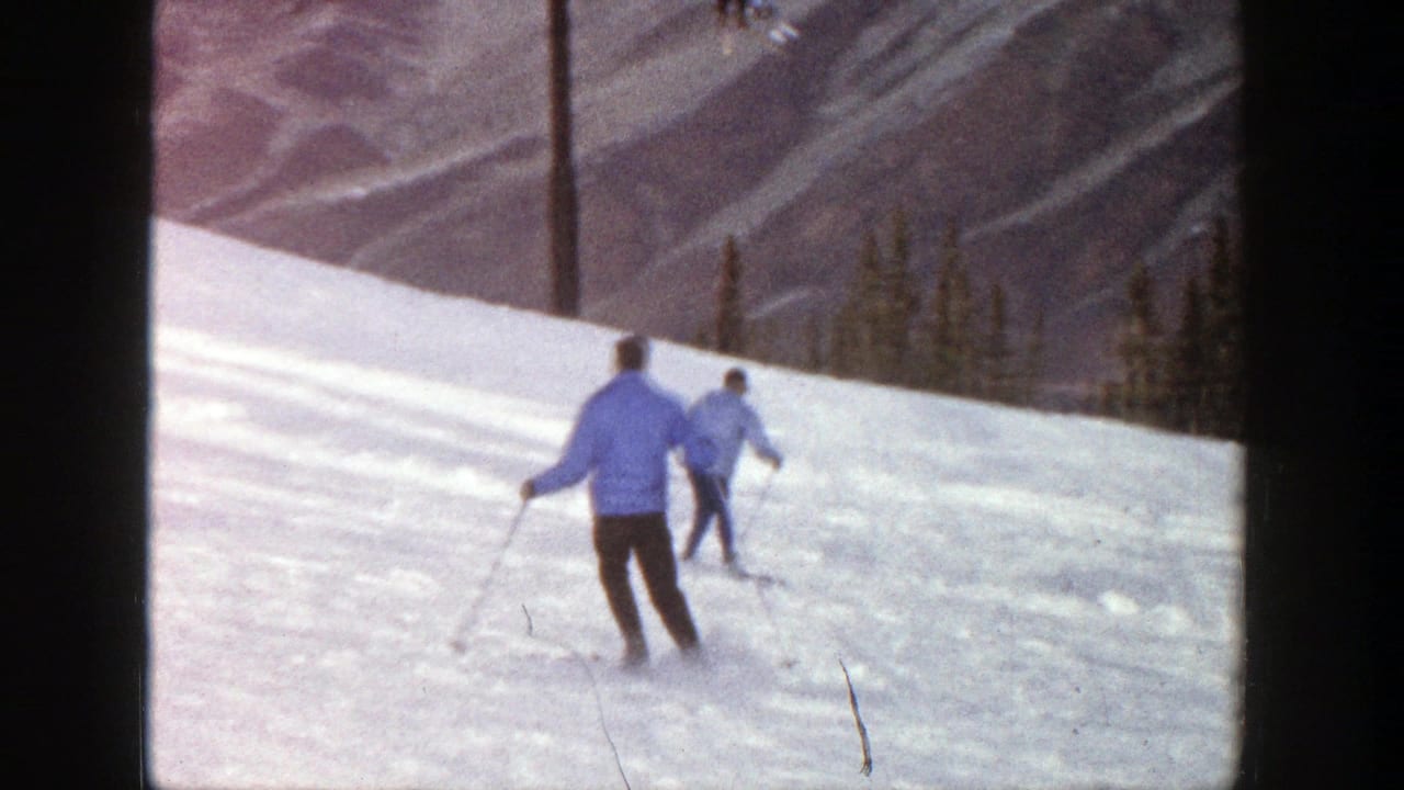 Two skiers continue downhill across bright snow near a mountain backdrop.