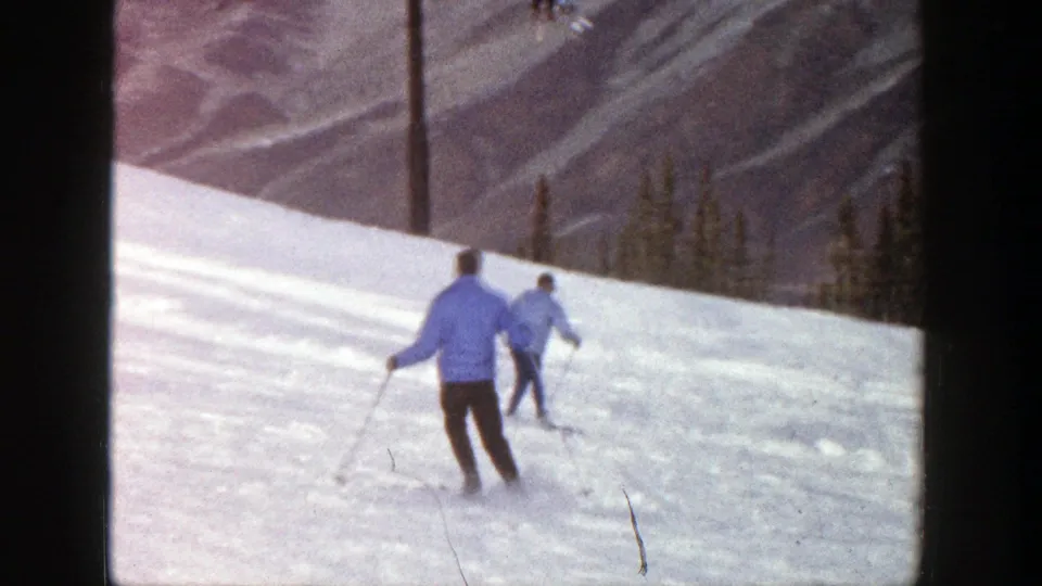1961 Aspen footage frame with two recreational skiers moving downhill on a wind-marked snowy slope.