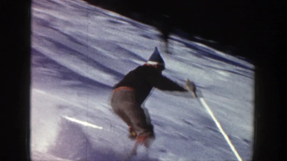 Adult skier in vintage Aspen footage leans across the snow while turning near a visible slope marker.