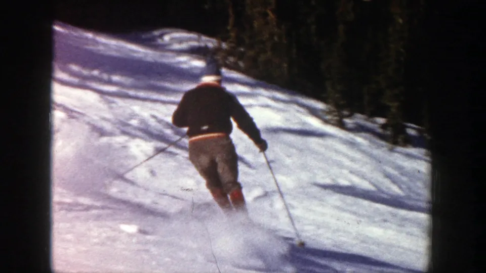 The Aspen skier is seen from behind, moving downhill across sunlit snow toward a darker tree line.
