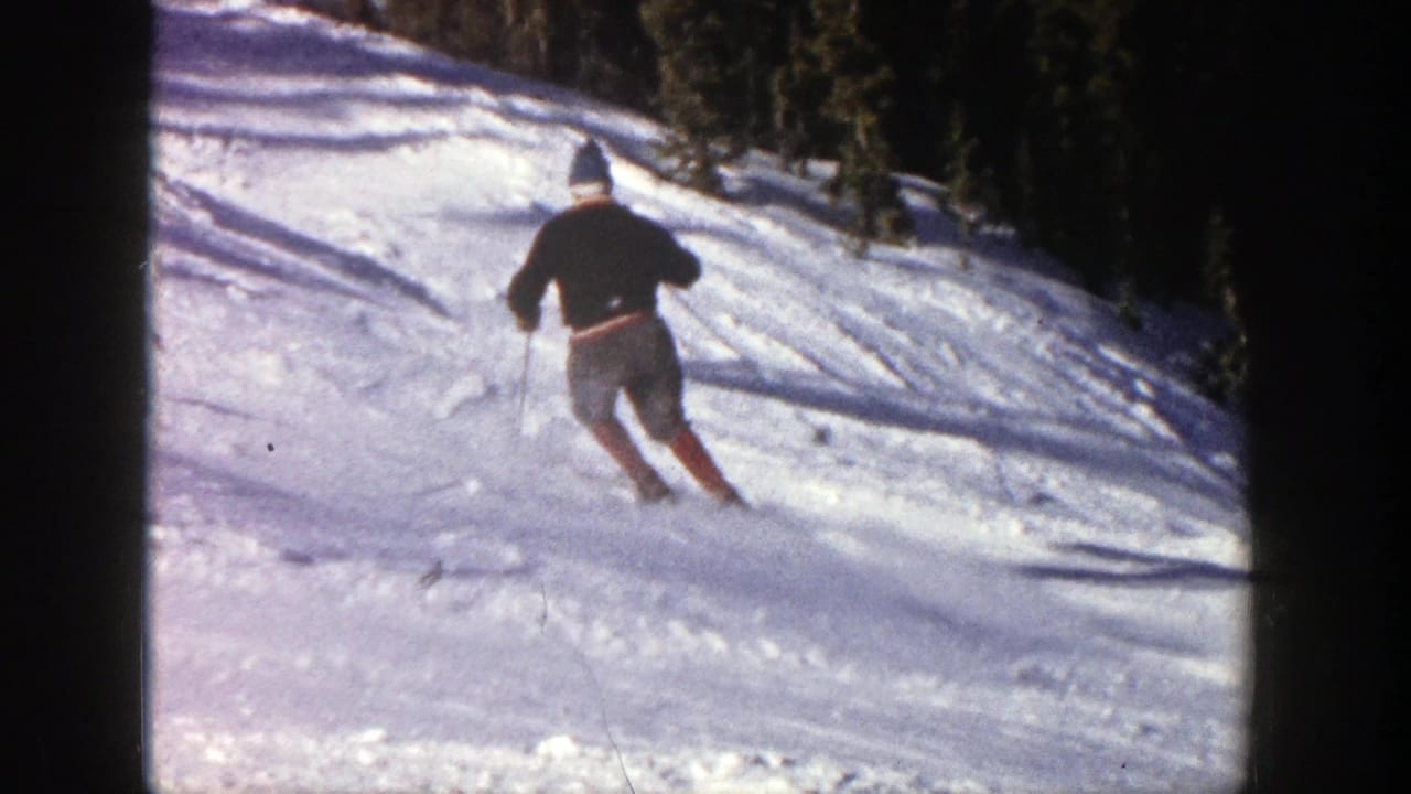 In Aspen, Colorado, a 1961 8mm frame shows a skier in dark outerwear carving across a sunlit snowy slope.