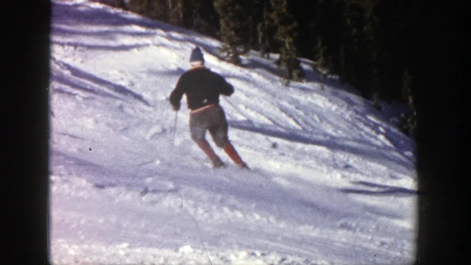 In Aspen, Colorado, a 1961 8mm frame shows a skier in dark outerwear carving across a sunlit snowy slope.