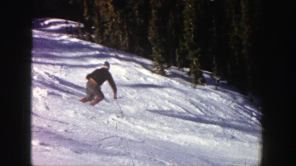 The skier leans through a downhill turn on the Aspen slope, with tree line and tracked snow visible behind.