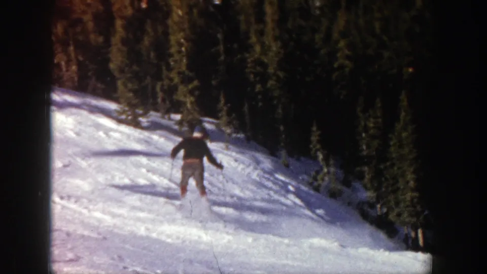 A distant skier continues down the snowy Aspen run, framed by dark evergreen trees and 8mm film edges.