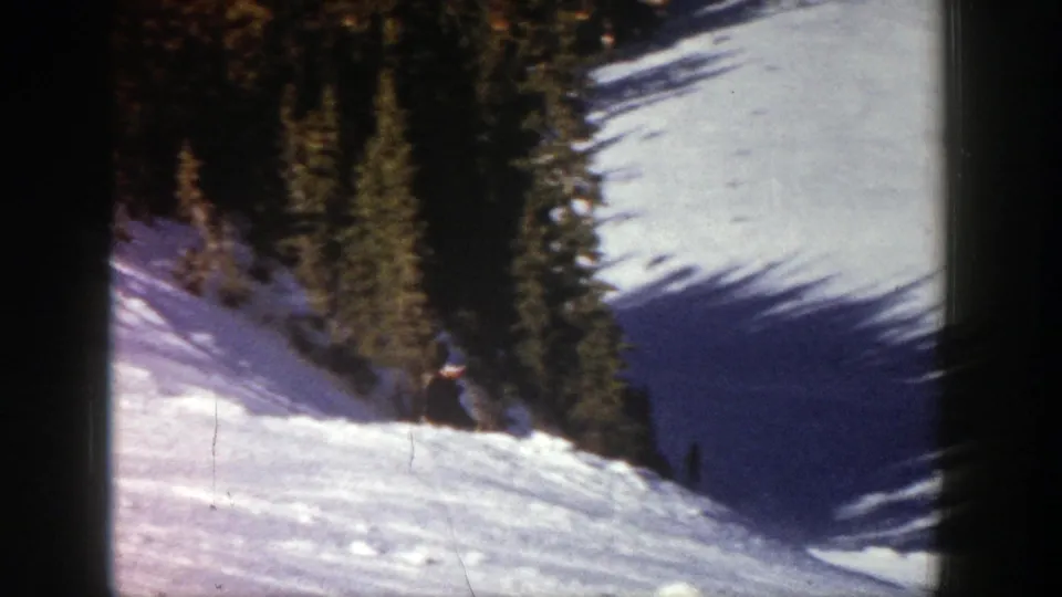 A distant skier in dark clothing descends a snowy Aspen slope in 1961, with evergreen trees along the run.