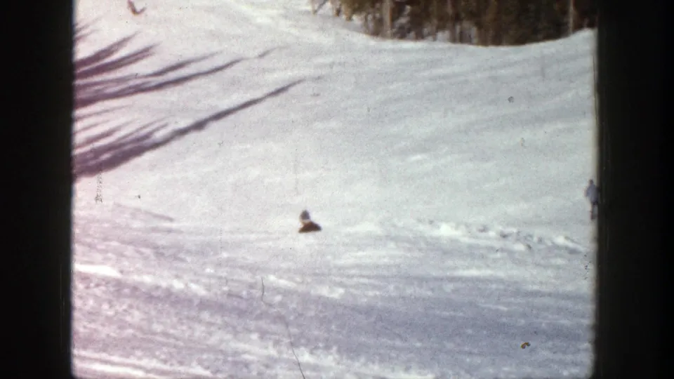 The skier appears small against a broad snowy Aspen slope, with trees and worn tracks visible in the distance.