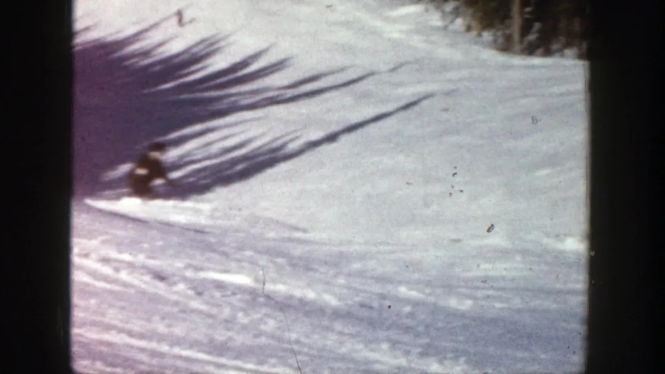 A skier in dark winter clothing crouches low while turning across a snowy Aspen slope in the 1961 home movie.