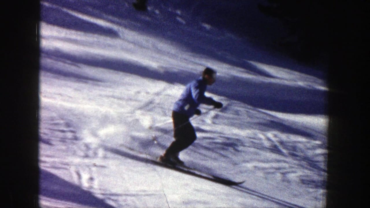 In Aspen, Colorado, 1961 footage, a skier in a blue jacket carves down a sunlit snowy slope with poles.