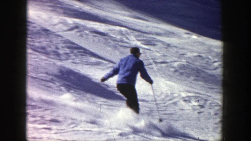 The skier in a blue jacket turns across the Aspen slope, with sunlit snow and tracks visible around them.