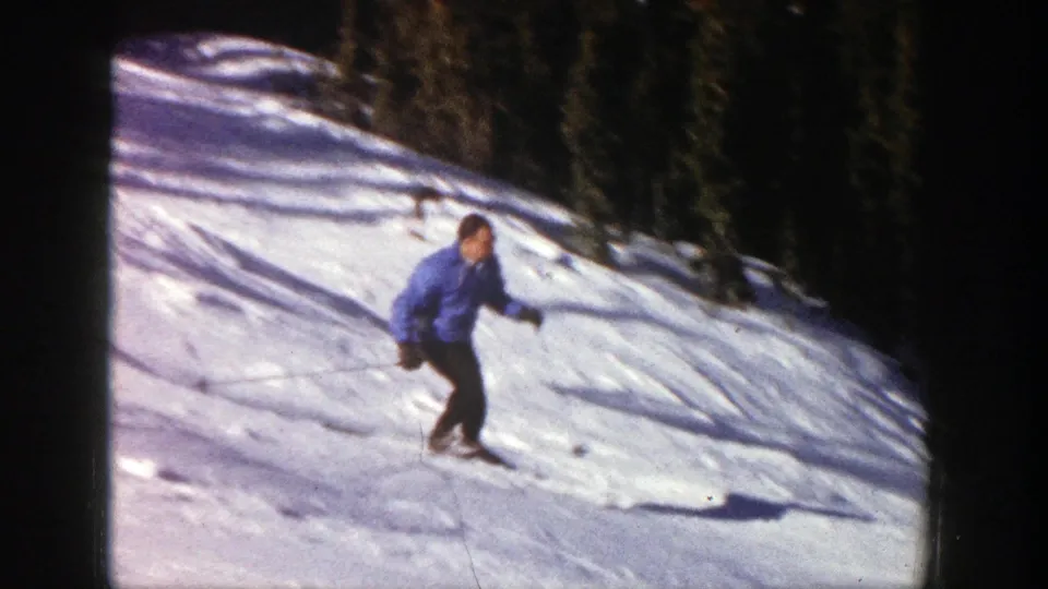 A man in a blue jacket skis through bright, tracked snow in the supplied Aspen, Colorado home-movie frame.