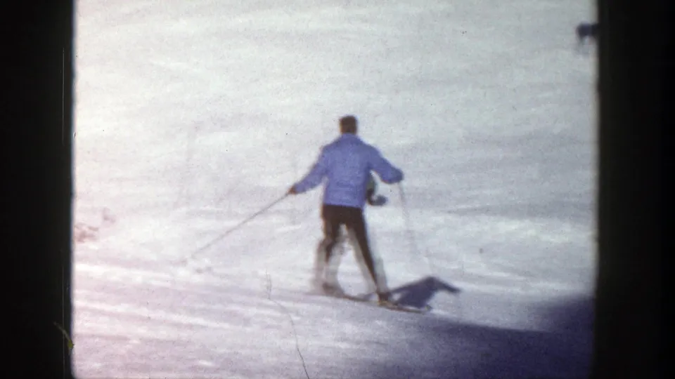 Adult skier in a blue sweater is seen from behind while descending a sunlit Aspen slope in 1961 footage.