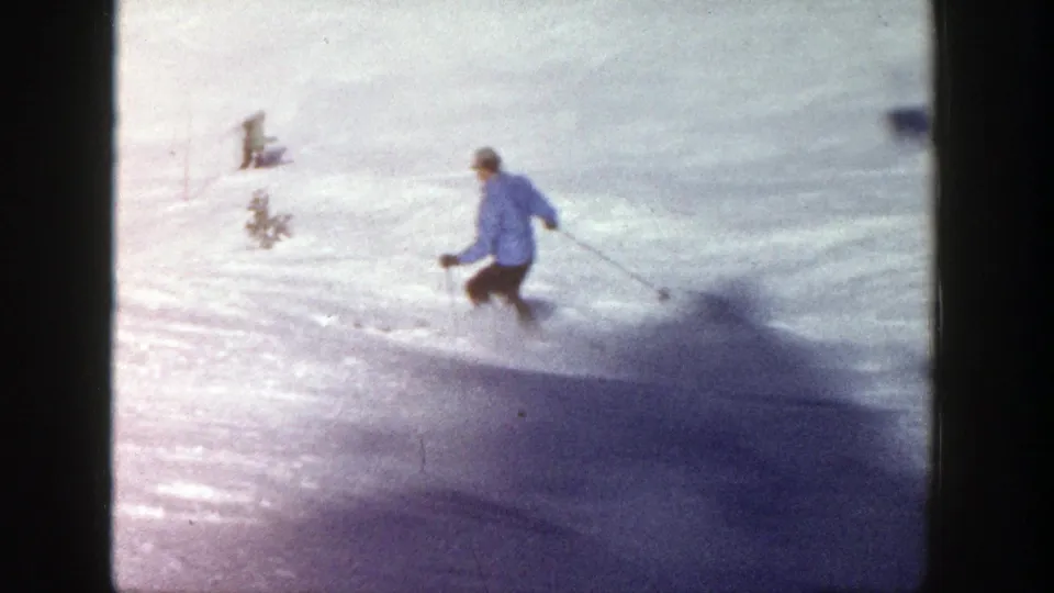 A skier in a blue sweater turns downhill near small trees on the sunlit Aspen slope in 1961 home-movie footage.