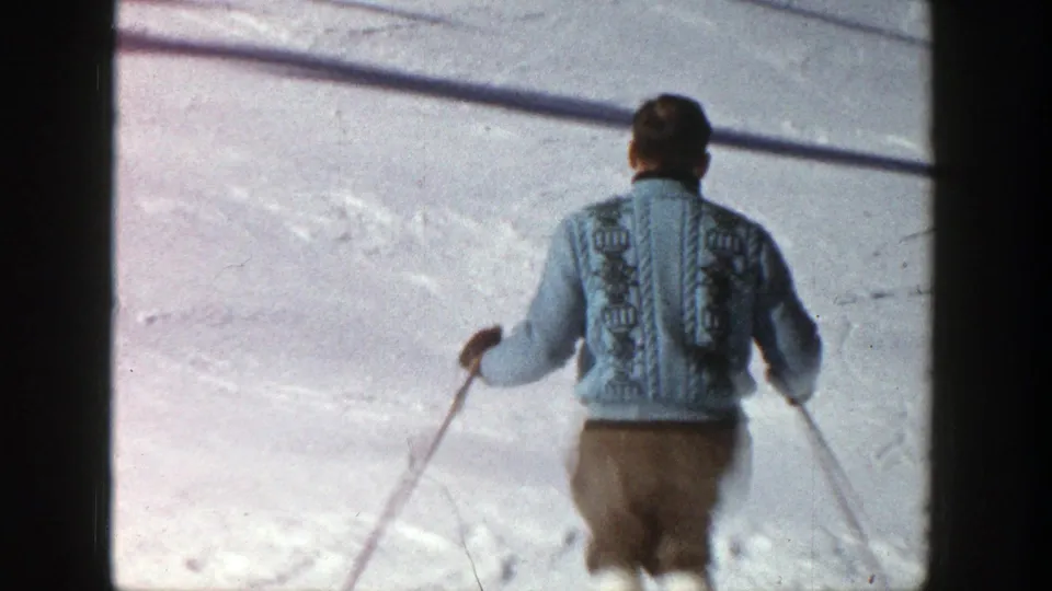 Adult man wearing a patterned blue sweater and brown knickerbockers skis away on a bright Aspen slope in 1961 footage.
