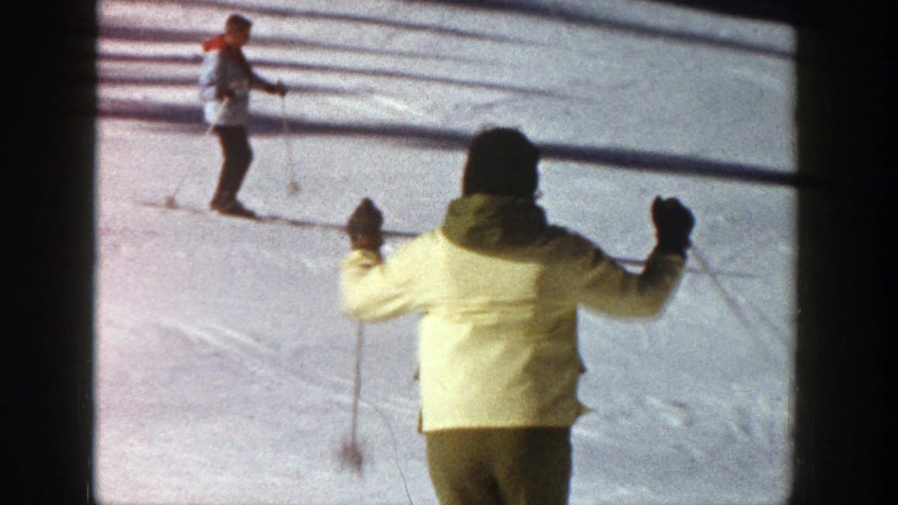 1961 Aspen home-movie frame showing a skier in a yellow jacket and dark pants moving downhill toward another skier on the snow.