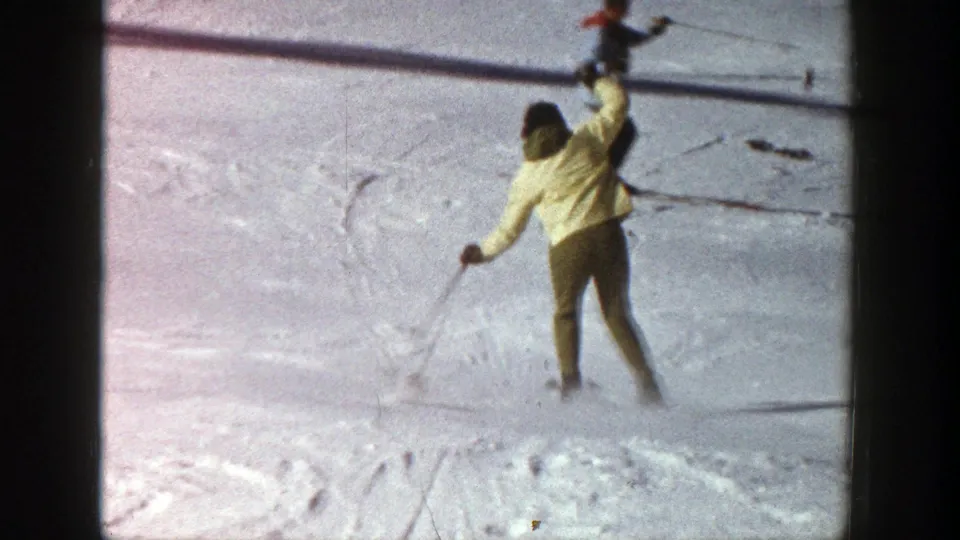 1961 Aspen skiing frame with a yellow-jacketed skier viewed from behind, one arm raised while moving across snow.