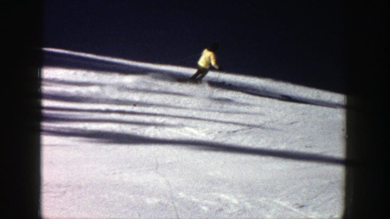 Distant skier in a yellow jacket crosses a snowy slope with long shadows.