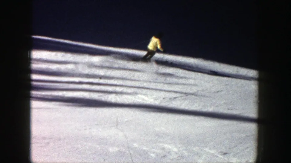 In Aspen, Colorado, 1961, a distant skier in a yellow jacket moves across a snowy slope with dark shadows on the snow.