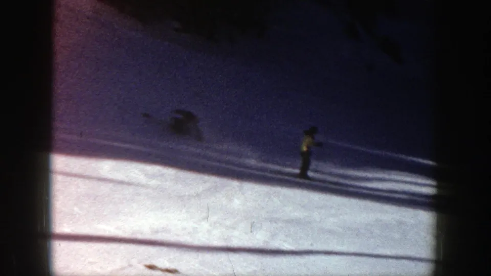 A skier in a yellow jacket is seen lower on the Aspen slope, with strong tree shadows and visible 8mm film texture across the frame.