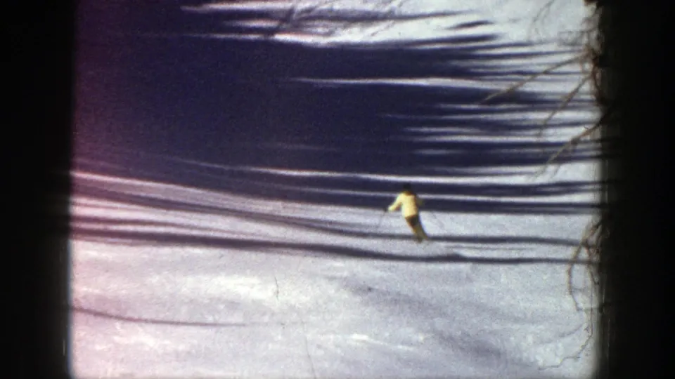 1961 Aspen home-movie frame showing a skier in a yellow jacket moving downhill across a snow-covered run with tree shadows.