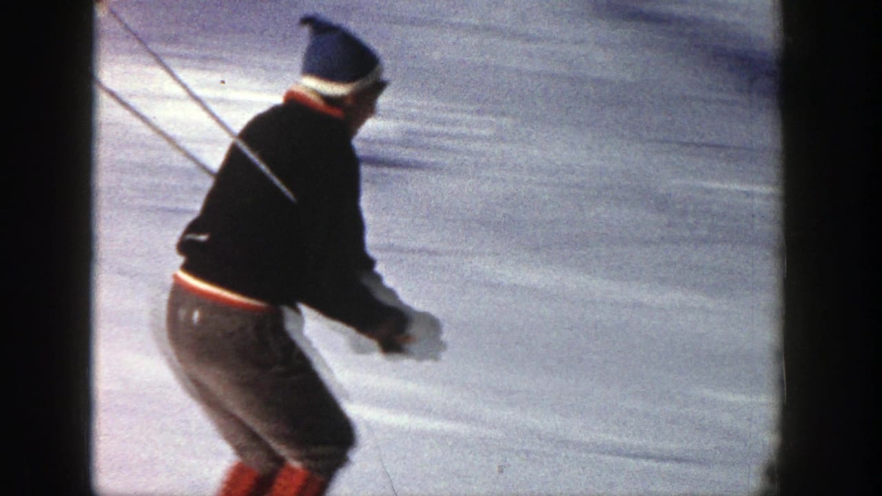 A skier in Aspen, Colorado, 1961, wearing a dark jacket, blue knit hat, and red-accented pants, moves across snow with poles.