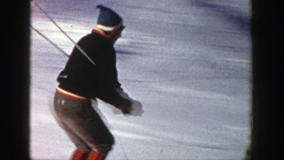 A skier in Aspen, Colorado, 1961, wearing a dark jacket, blue knit hat, and red-accented pants, moves across snow with poles.