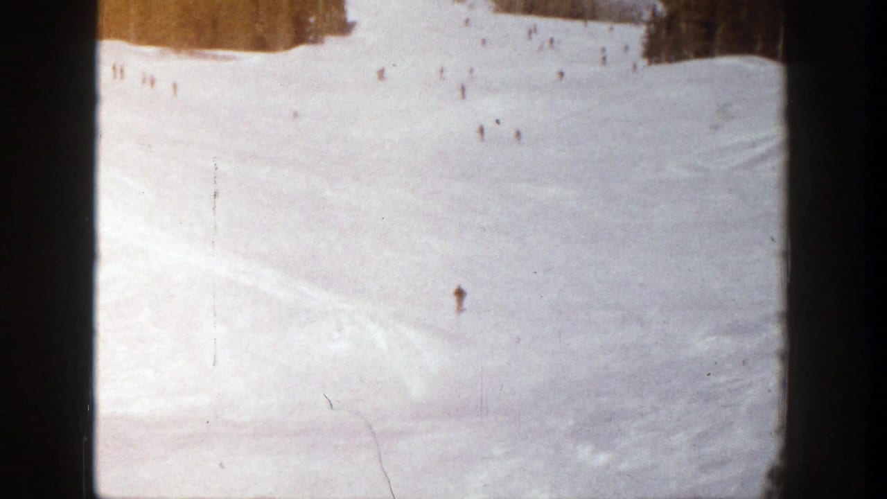 1961 Aspen home-movie frame showing small skiers descending a wide snow-covered slope with trees near the top edge.