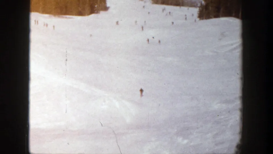 1961 Aspen home-movie frame showing small skiers descending a wide snow-covered slope with trees near the top edge.