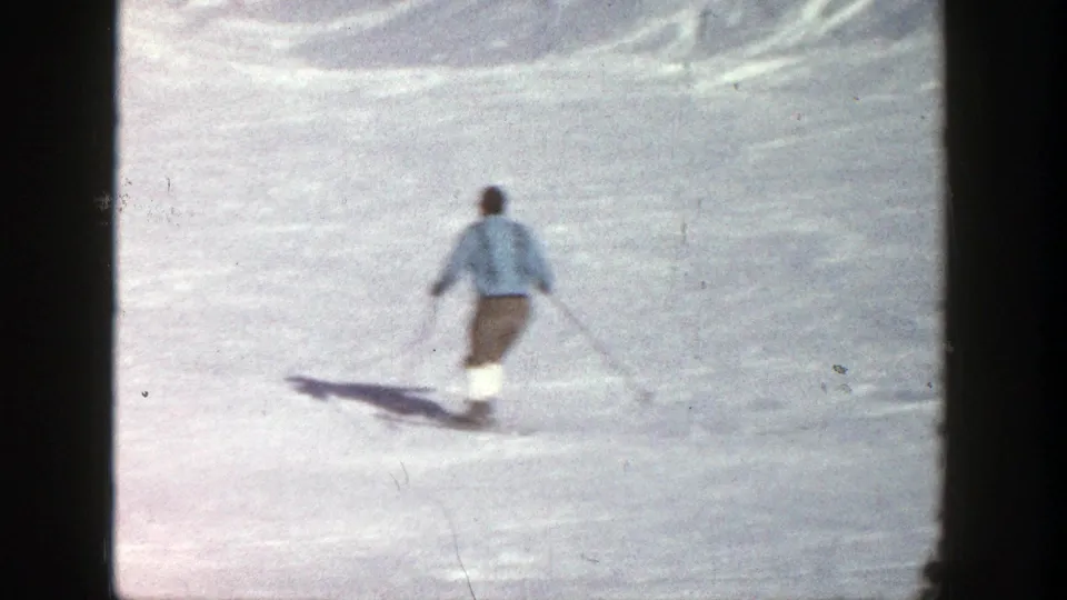 A skier in a light blue jacket and brown pants is filmed from behind on an Aspen snow slope in 1961 footage.