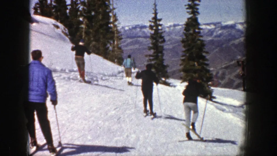 Several skiers move along an Aspen slope in 1961, with evergreen trees, distant mountain ridges, and strong sunlight visible.