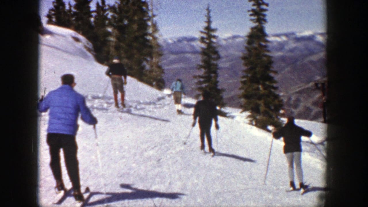 1961 Aspen home-movie frame showing skiers practicing on a sunny snow slope with evergreens and mountain ridges beyond.