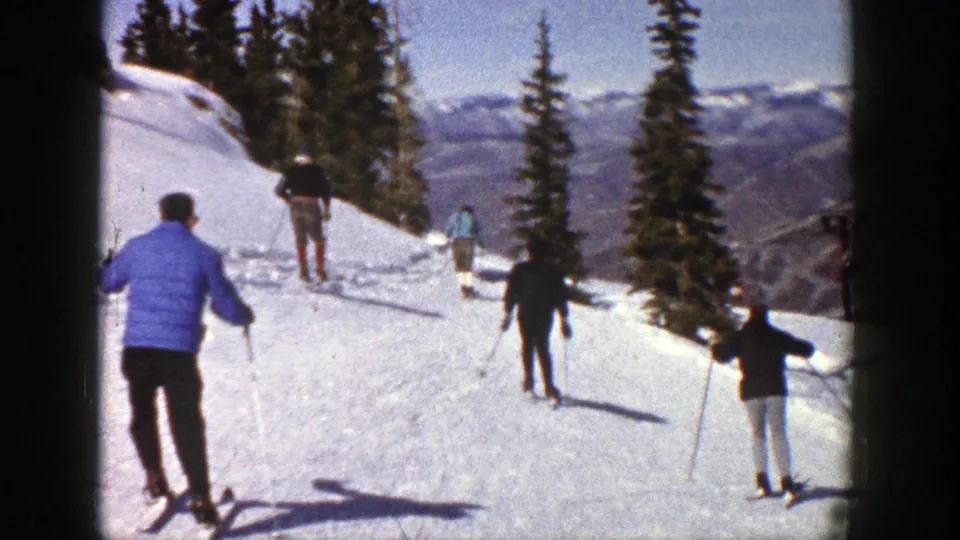 1961 Aspen home-movie frame showing skiers practicing on a sunny snow slope with evergreens and mountain ridges beyond.