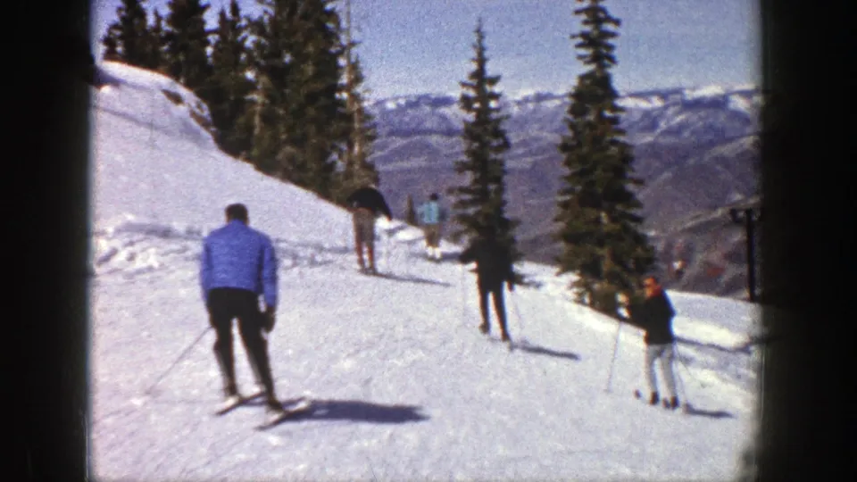 1961 Aspen skiing footage frame showing a small group gliding down a snow-covered trail with evergreens and mountains visible.