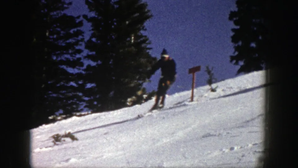Aspen home-movie frame from 1961 showing one skier near a small sign on a bright snow slope beside evergreens.
