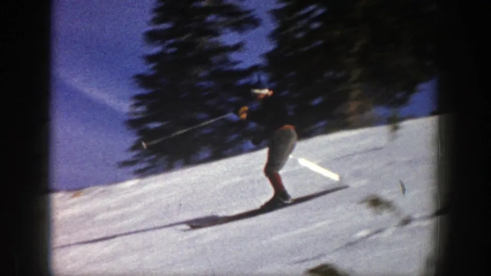 A skier in a dark top and red socks descends an Aspen snow slope with long shadows and evergreen trees behind him.
