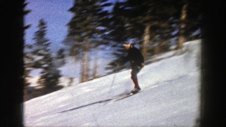The 1961 Aspen home-movie frame shows a skier crossing the hillside, softened by motion blur and 8mm grain.