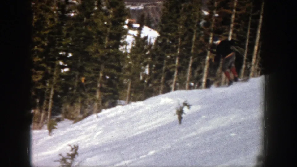 A skier appears on the right side of a snowy Aspen slope, with dense evergreens and soft 8mm exposure in the background.