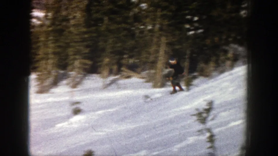 The 1961 Aspen home-movie frame shows a skier crossing a snowy run, partly blurred by motion and surrounded by evergreen trees.