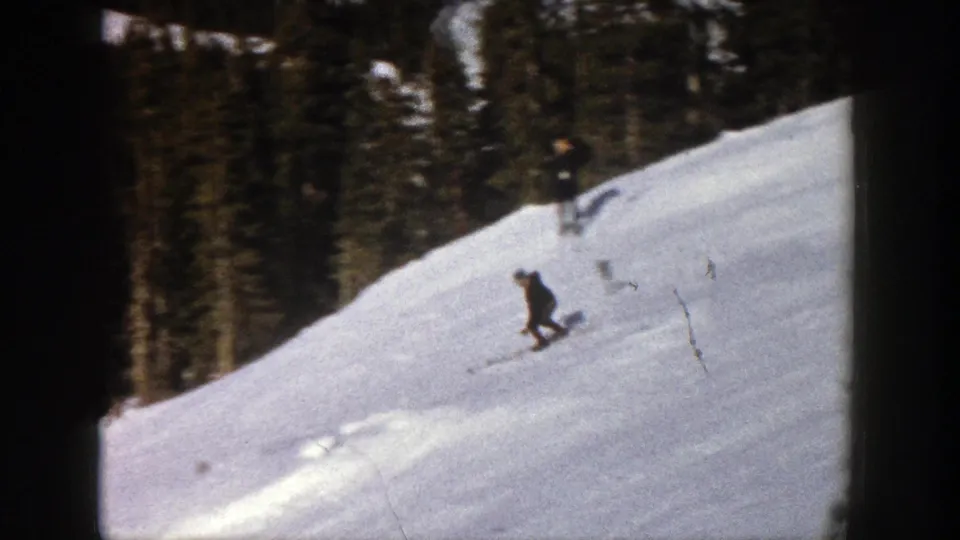 A distant skier in dark clothing turns on a snowy Aspen slope, framed by dense evergreens.