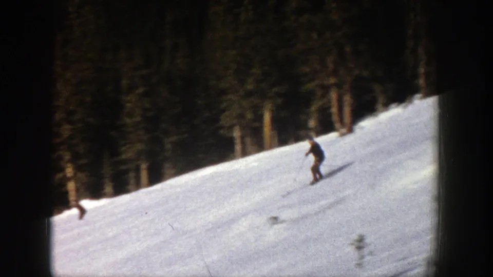 1961 Aspen ski footage shows a skier moving across the hillside in front of a dark forest edge.