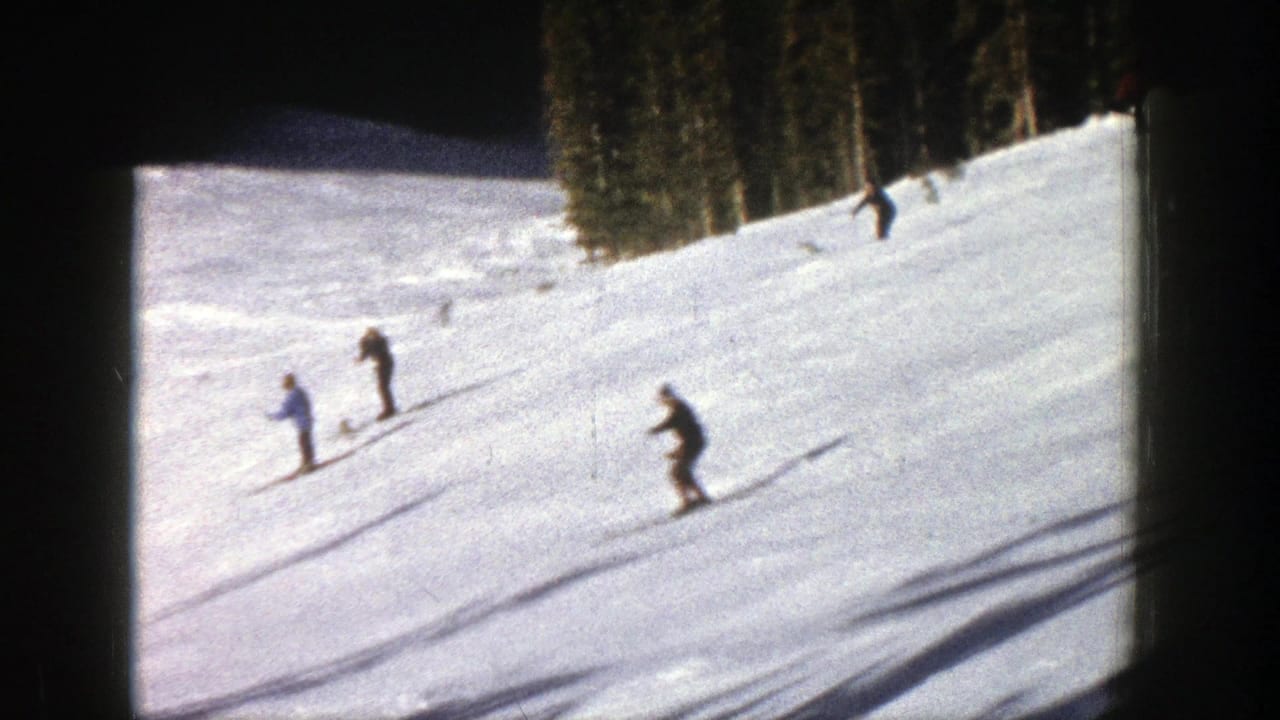 In 1961 Aspen footage, distant skiers descend a bright snowy mountain slope with trees along the upper edge.