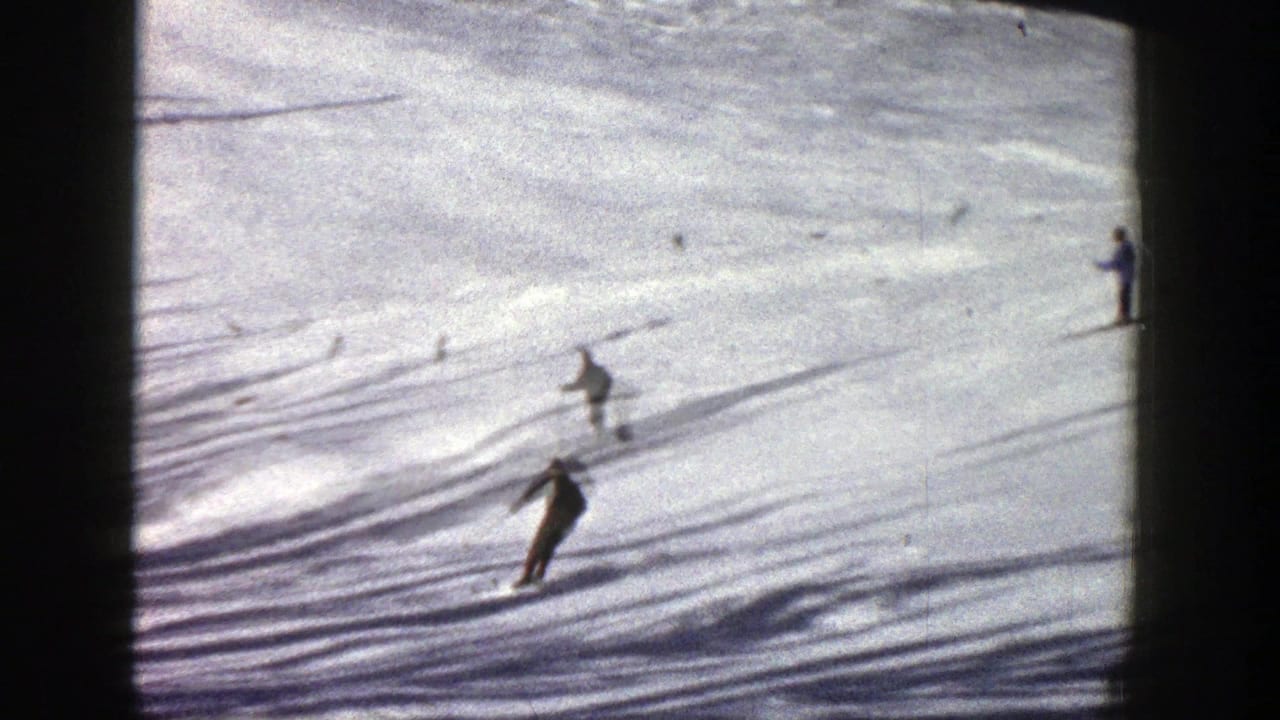 Skiers descend a bright snowfield with long shadows across the surface.
