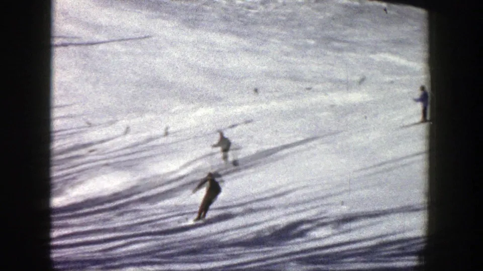 Distant skiers in the Aspen home-movie clip move downhill across a sunlit snowfield with pronounced shadows.