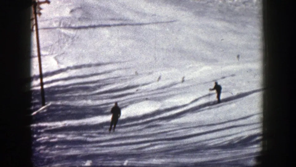 Two distant skiers move across an Aspen ski slope, with tracks and a vertical pole or tree trunk near the frame edge.