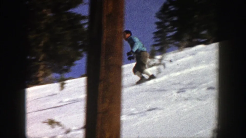 In Aspen, Colorado in 1961, a man in a blue sweater skis across a snowy mountain slope framed by dark evergreens.