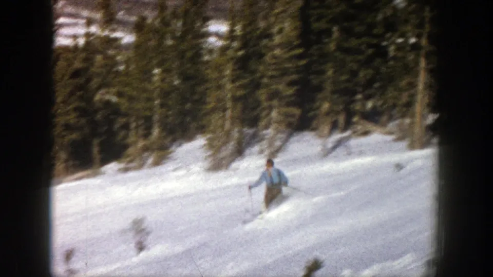 The Aspen skier moves downhill through bright snow, filmed from a handheld 8mm viewpoint with trees behind him.