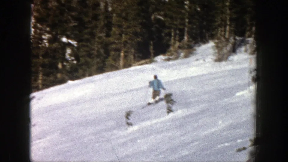 Near the end of the 1961 Aspen clip, the skier descends past low brush on a snowy slope below evergreen trees.