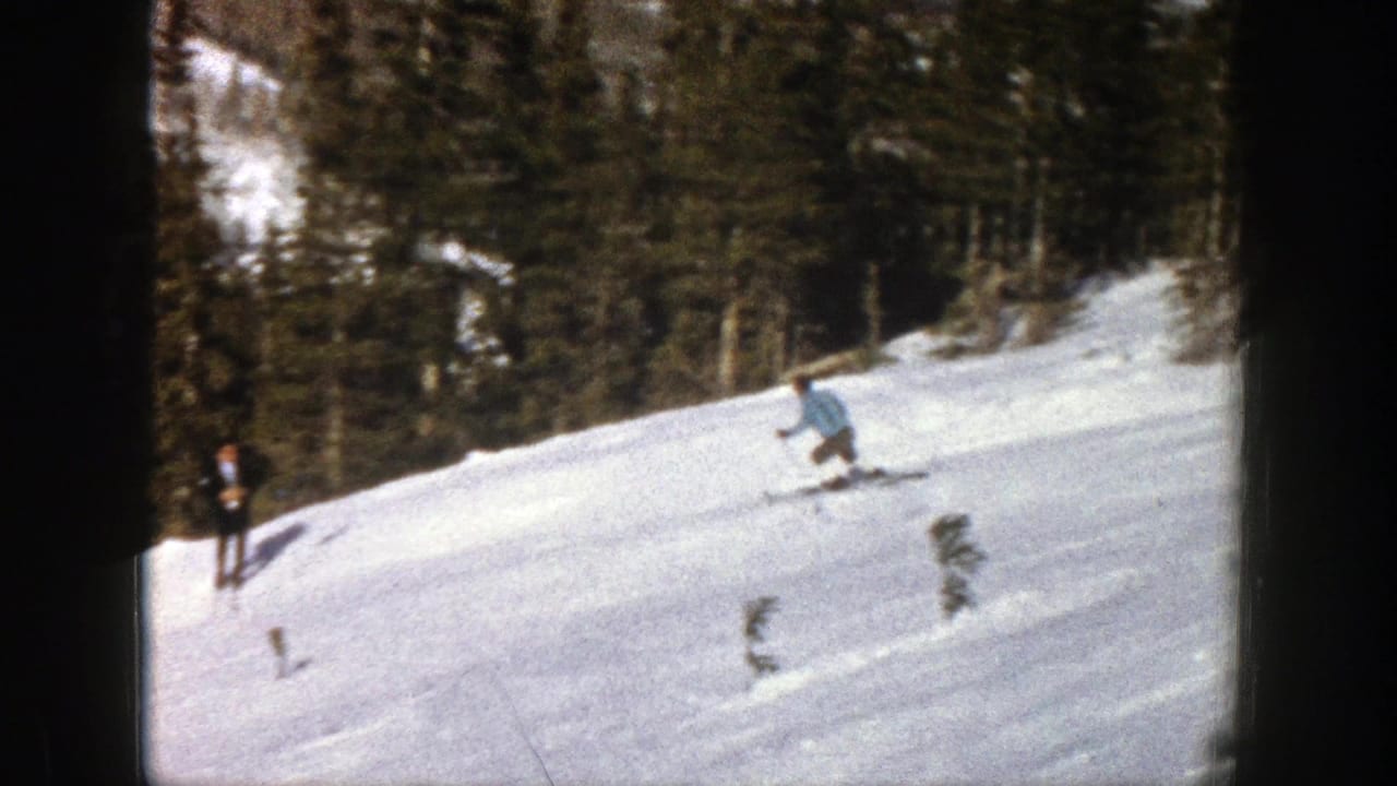 In Aspen, Colorado, 1961, a skier in a light blue jacket moves downhill while another skier stands near the left edge of the wooded slope.
