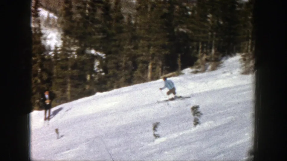 In Aspen, Colorado, 1961, a skier in a light blue jacket moves downhill while another skier stands near the left edge of the wooded slope.