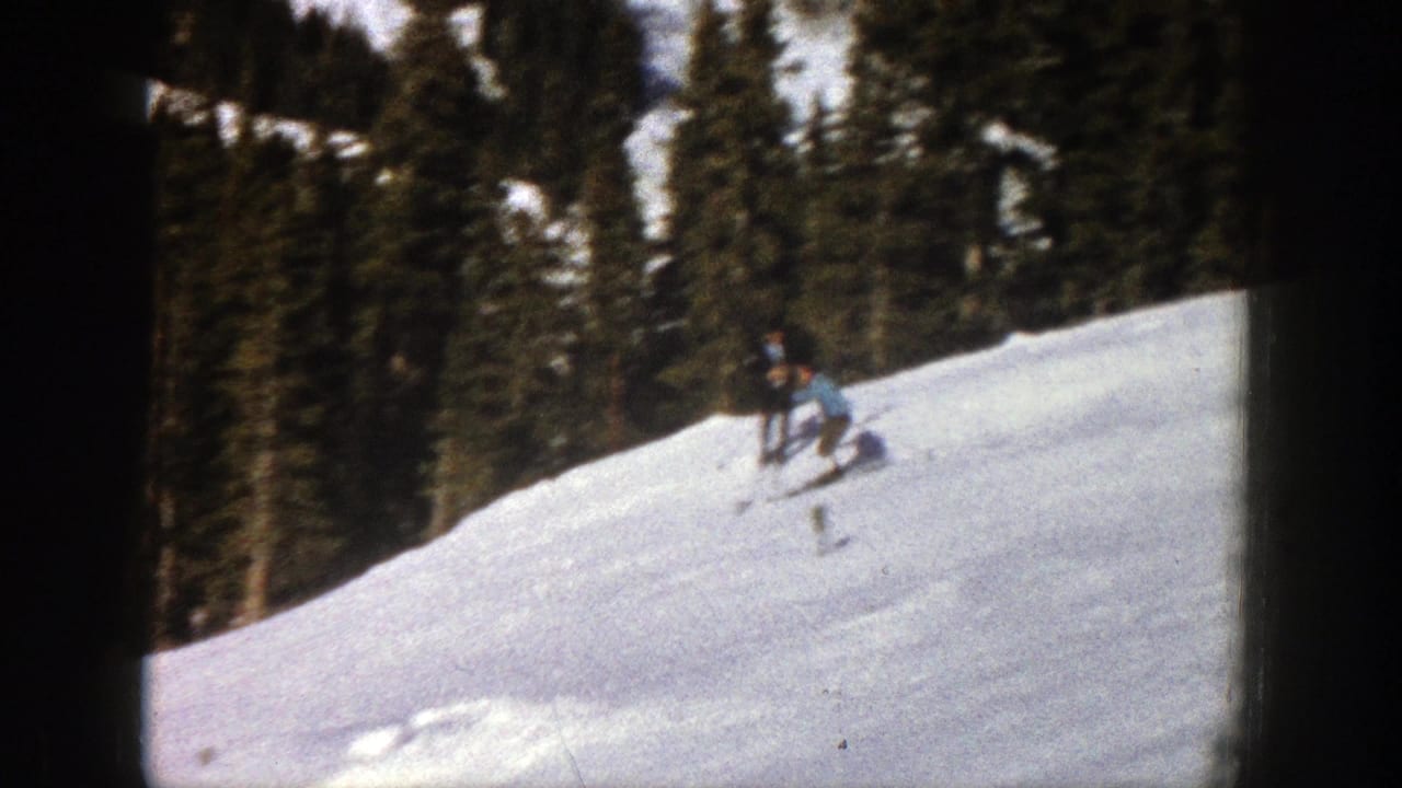 Skier in blue carves across a snowy hillside with trees behind.